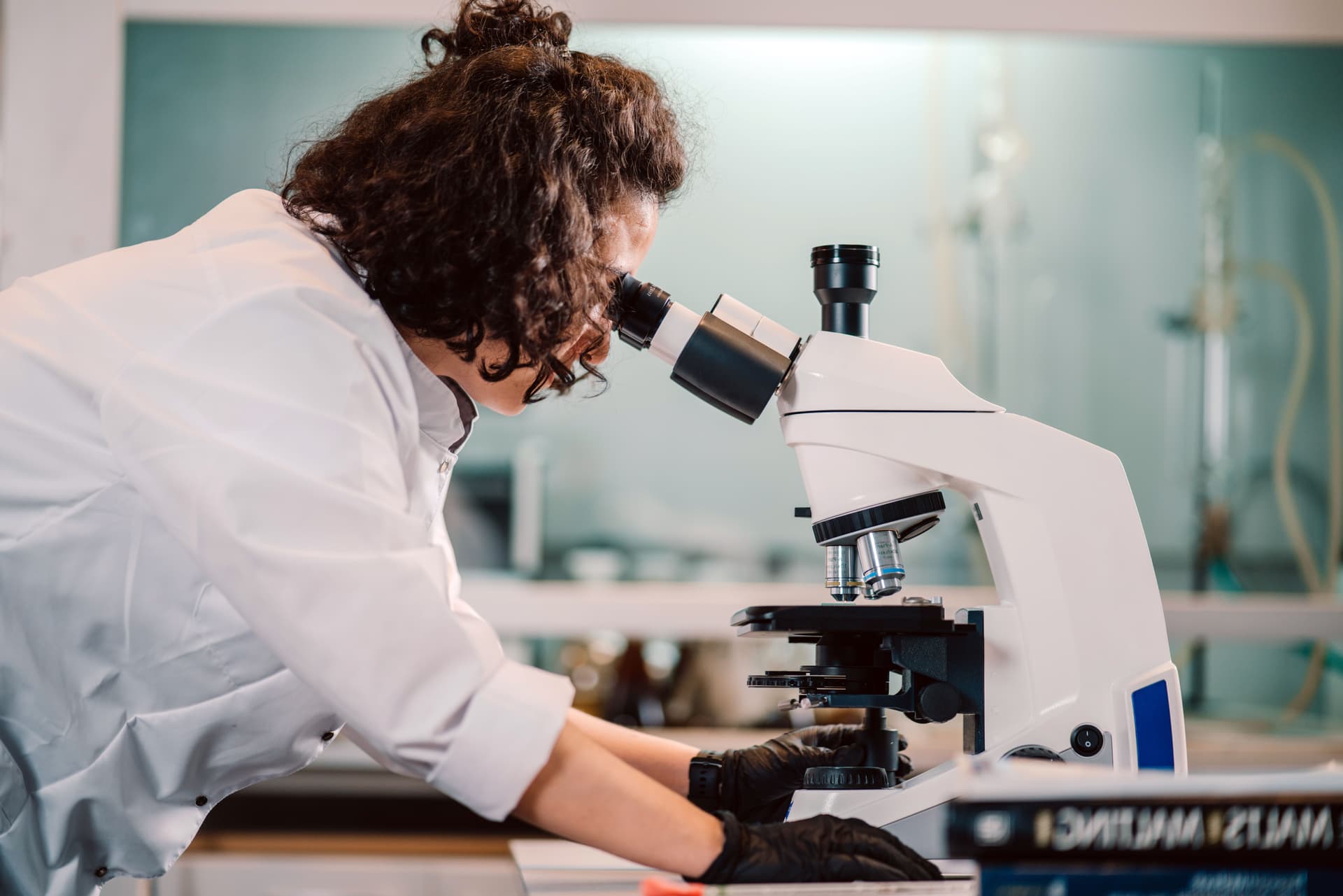 A scientist in a white lab coat uses a microscope in a laboratory setting, with laboratory equipment visible in the background.